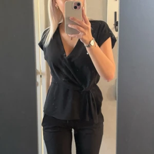Svart omlottopp från Zara - Snygg svart omlottopp från Zara med v-ringning och knyt i midjan. Toppen har korta ärmar och en stilren, feminin passform som framhäver midjan. Perfekt för dig som gillar en enkel men trendig look. Storlek XS