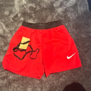 Röda Nike Dri-FIT träningsshorts M - Snygga röda träningsshorts från Nike med svart resår i midjan och vit swoosh-logga på benet. Shortsen har snörning i midjan och reflekterande detaljer baktill. Tillverkade i lätt och snabbtorkande Dri-FIT polyester, perfekta för intensiva träningspass.