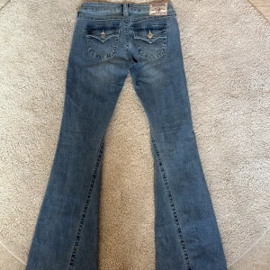 Blå bootcut jeans från True Religion - Säljer ett par klassiska blå bootcut jeans från True Religion med snygga fickdetaljer och logga bak. Jeansen har låg midja och utsvängda ben, samt dekorativa sömmar och knappar på bakfickorna.