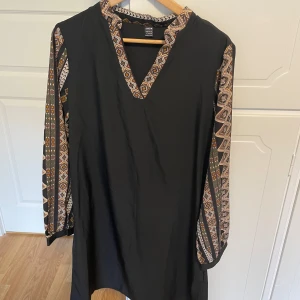 Svart mönstrad klänning från SHEIN - Svart klänning från SHEIN i storlek L med långa ärmar och färgglada etno-inspirerade mönster längs ärmar och v-ringad hals. Rak passform och mjukt, lätt material. Perfekt för dig som gillar unika detaljer och bekväm stil.