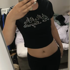 Svart croppad topp med strasstext - Svart croppad topp med kort ärm och glittrande strasstenar framtill som bildar texten 'No Regrets' med flammor. T-shirten har en tight passform och är perfekt för dig som gillar edgy och trendiga detaljer.
