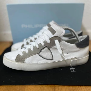 Vita sneakers från Philippe Model - Snygga vita sneakers från Philippe Model med grå mockadetaljer och vit snörning. Skorna har en rund tå, platt sula och diskret logga på sidan. Ovandelen är i skinn med kontrasterande grå hälkappa och detaljer längs snörningen. Perfekta för en clean och stilren look. Bra skick! Storlek 44. Allt org medföljer! Skriv för frågor! 💬 Nypris runt 3000 kr!