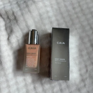 CAIA Dewy Drops Serum Foundation 10W - CAIA Dewy Drops Serum Foundation i nyansen 10W. Kommer i en snygg frostad glasflaska med silvrig pump och tillhörande metallic-förpackning. Ger en naturligt glowig finish och innehåller vitamin E och glycerin för extra fukt. Perfekt för dig som vill ha en fräsch och återfuktad look.
