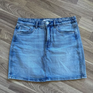 Blå jeanskjol från Kappahl - Snygg blå jeanskjol från Kappahl i klassisk modell med fem fickor, bälteshällor och dragkedja med knapp framtill. Kjolen är kort och har en lätt tvättad look som ger en cool vibe. Perfekt att matcha med sneakers eller boots.