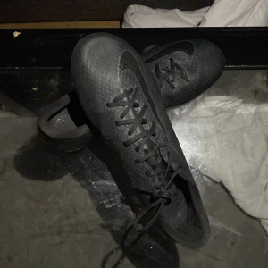 Svarta Nike fotbollsskor med snörning ST 45 använda - Säljer ett par svarta Nike fotbollsskor med snyggt mönstrad ovandel och klassisk snörning. Skorna har rund tå och dobbar för bra grepp på planen. Perfekta för dig som vill ha stilrena och funktionella skor till match eller träning.