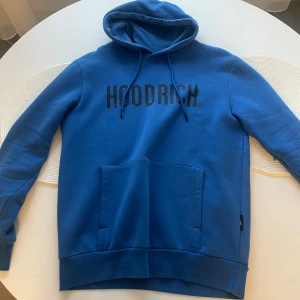 Blå hoodie från Hoodrich med tryck - Snygg blå hoodie från Hoodrich med svart logotyptryck över bröstet. Tröjan har huva med snörning och en stor magficka framtill. Perfekt för dig som gillar streetwear och vill ha en bekväm och stilren look.