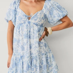 Blommig ljusblå kortklänning - Supersöt kortklänning med ljusblått blommönster på vit botten. Klänningen har puffiga kortärmar, knytning vid bysten och volangig kjol. Helt ny, nypris 500