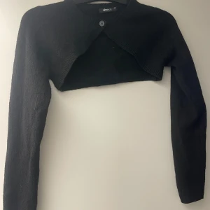 Svart croppad kofta från Gina Tricot - Svart croppad kofta från Gina Tricot med lång ärm och en knapp framtill. Koftan är stickad och har en enkel, stilren design som passar perfekt över en topp eller klänning. Perfekt för lager-på-lager-stil under kyliga dagar.