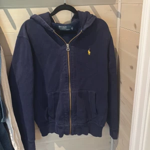 Mörkblå hoodie från Polo Ralph Lauren - Snygg mörkblå hoodie från Polo Ralph Lauren med gul broderad logga på bröstet. Tröjan har dragkedja, huva med strukturmönster på insidan och två stora fickor framtill. Perfekt för en chill och stilren look.