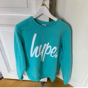 Turkos sweatshirt från Hype - Turkos sweatshirt från Hype med stort vitt tryck framtill. Tröjan har rund hals, långa ärmar och är tillverkad i en mjuk bomulls- och polyestermix. Perfekt för dig som gillar streetstyle och vill sticka ut med en färgglad look.