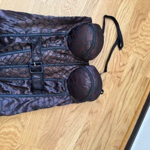 Brun leopardmönstrad korsett med bälte - Snygg brun korsett med leopardmönster och svarta detaljer. Toppen har vadderade kupor, avtagbara axelband och ett dekorativt bälte framtill. Materialet är glansigt och ger en lyxig känsla. Perfekt för dig som vill sticka ut med en edgy och trendig look.