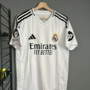 Real Madrid Bellingham 5 matchtröja - Snygg vit Real Madrid fotbollströja från Adidas med Bellingham och nummer 5 på ryggen. Tröjan har svarta detaljer, klubbmärke, sponsortryck och Champions League-märke på ärmen. Klassisk V-ringning och korta ärmar i lätt, ventilerande material. Helt ny med tagg en replika