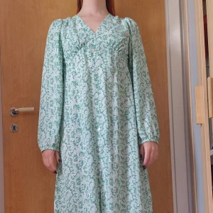 Grönblommig långklänning med v-ringning - Blommig grön klänning från Escape. Långa ärmar, markerad midja, luftigt skön kjol och dekorationsknappar framtill. Liten strl L men skulle även passa M. Endast använd 1 gång, mycket fint skick!  Katt i hemmet.  