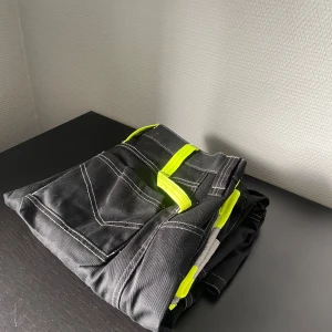 Svarta cargopants med reflex och neon - Svarta cargopants med vita sömmar, flera fickor och detaljer i neon-gult samt reflexband vid benen. Byxorna har en loose fit och är perfekta för dig som vill sticka ut. Materialet är slitstark bomull och polyester, perfekt för aktiva dagar.
