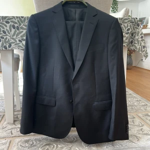 Svart kostym Dressmann slim fit ull - Säljer en klassisk svart kostym från Dressmann i slim fit-modell. Kostymen är tillverkad i 100% ull och består av kavaj och matchande byxor. Snygg passform med två knappar framtill, fickor med lock och slät design utan mönster. Perfekt för dig som vill ha en stilren look. Den har varit använd bara en gång. 