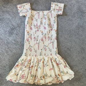 Blommig kortklänning med volang - Supersöt kortklänning med blommigt mönster i ljusa färger. Klänningen har fyrkantig halsringning, korta ärmar och en volangig kjol. Perfekt för sommarens alla tillfällen och ger en romantisk vibe.