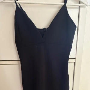 Svart tight kortklänning med v-ringning - Snygg svart klänning med smala axelband och v-ringad hals. Modellen är figurnära och kort, med dragkedja i ryggen för enkel påklädning. Perfekt för dig som gillar stilrena och minimalistiska plagg med en modern siluett.