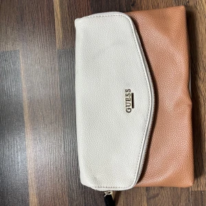 Beige och brun kuvertväska från Guess - Snygg kuvertväska från Guess i beige och brun med struktur i materialet. Väskan har ett stilrent metallogo framtill, dragkedja och magnetknapp. Insidan är ljus med en liten innerficka med dragkedja. Perfekt för att bära det viktigaste med stil.