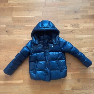 Jacka Polo Ralph Lauren - Snygg blå dunjacka från Polo Ralph Lauren med huva och broderad logga på bröstet. Jackan är quiltad och har dragkedja framtill samt två fickor. Perfekt för kalla dagar och ger en cool look med sin blanka finish. Priset går att diskutera😊