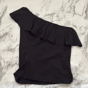 Svart oneshoulder volangtop - Snygg svart topp med oneshoulder-design och bred volang längs halsringningen. Toppens stilrena look gör den lätt att matcha och den har en figurnära passform. Perfekt för dig som vill sticka ut med en trendig och modern vibe. Mammas gamla ❤️