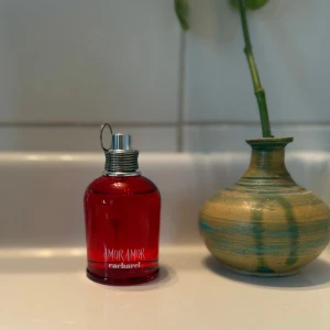 Amor Amor parfym från Cacharel - Säljer en Amor Amor parfym från Cacharel. Flaskan är klarröd med silvrig metallkork och har en rund, kompakt form. Perfekt för dig som gillar fruktiga och blommiga dofter med en ungdomlig vibe. Flaskan är tillverkad av glas och har en snygg, modern design.