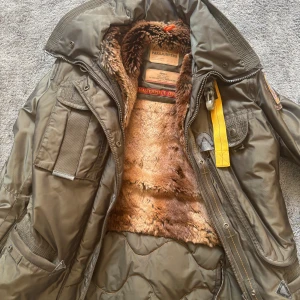 Parajumpers brun parkajacka  - Parkajacka från Parajumpers i mörkbrun färg med fluffigt pälsfoder på insidan. Jackan har flera fickor, gul detaljrem och broderad patch på ärmen. Yttermaterialet är slitstark polyester och jackan stängs med dragkedja och knappar. Perfekt för kalla dagar.