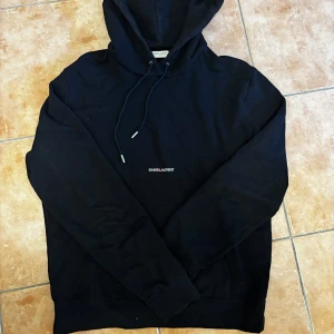 Svart hoodie från Saint Laurent - Säljer en svart hoodie från Saint Laurent med minimalistisk logga på bröstet. Klassisk modell med huva och snörning, tillverkad i mjuk bomull. Perfekt för dig som gillar stilrena och exklusiva plagg med streetkänsla.