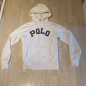 Vit hoodie från Polo Ralph Lauren - Snygg vit hoodie från Polo Ralph Lauren med stor broderad POLO-text i mörkblått på bröstet. Klassisk känguruficka framtill, huva med snörning och långa  ärmar. Perfekt för en chill och stilren look. st m bara fråga om du har funderinga😁
