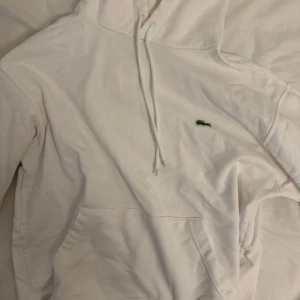 Vit hoodie från Lacoste med logga - Säljer en clean vit hoodie från Lacoste i klassisk passform. Tröjan har huva med snörning, känguruficka och den ikoniska gröna krokodilloggan broderad på bröstet. Perfekt för dig som gillar stilren och enkel design.