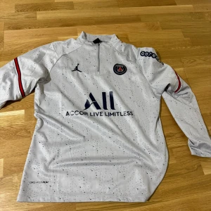 PSG långärmad träningströja Nike - Snygg långärmad PSG träningströja från Nike i ljusgrått med svarta prickar. Tröjan har halv dragkedja, PSG-logga på bröstet och Jordan-logga. Röd, vit och svart detalj på ärmarna. Materialet är lätt och andas, perfekt för fotbollsträning.