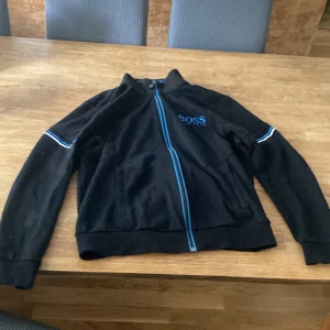 Svart ziptröja från Hugo Boss - Svart långärmad ziptröja från Hugo Boss med blå detaljer och logga på bröstet. Tröjan har blå dragkedja och vita/blå ränder på ärmarna. Perfekt för dig som gillar stilren och sportig look.