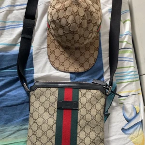 Gucci axelväska med GG-mönster - Säljer en snygg axelväska från Gucci med klassiskt GG-mönster i beige och brunt. Väskan har bred svart axelrem, detaljer i svart skinn och en ikonisk grön-röd rand i mitten. Perfekt för dig som vill ha en stilren och lyxig accessoar.