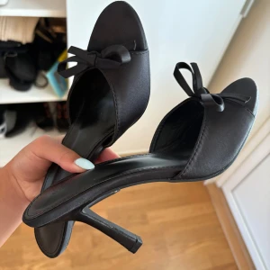 Svarta klackar med rosett o - Snygga svarta pumps med öppen tå och elegant rosett framtill. Skorna har en smal hög klack och är tillverkade i ett satinliknande material som ger en lyxig känsla. Perfekta för dig som vill ha en chic och stilren look med lite extra attityd.