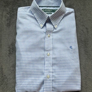 Blårutig skjorta från Ralph Lauren - Snygg fitted skjorta från Ralph Lauren med stretch och non iron-material. Skjortan har ett klassiskt rutigt mönster i blått och vitt, button-down krage och broderad RL-logga på bröstet. Perfekt för dig som gillar stilrena och fräscha plagg.
