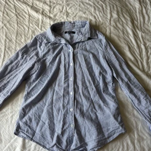 Skjorta - Snygg skjorta från Gina Tricot i vitt och blått med klassiskt randigt mönster. Skjortan har lång ärm, knappar framtill och en avslappnad krage. 