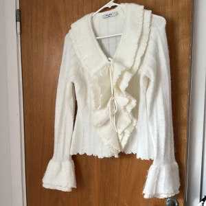 Vit kofta med volang från NA-KD - Supersöt vit kofta från NA-KD i storlek XS. Den har stora volanger vid kragen och ärmsluten, samt snörning framtill. Materialet är mjukt och stickat, perfekt för dig som gillar romantiska detaljer och vill ha något extra till din outfit.