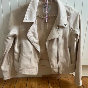 Beige mockajacka med dragkedjor - Snygg beige mockajacka från Lindex med klassisk bikerkrage och silvriga dragkedjor. Jackan har långa ärmar, två fickor med dragkedja och coola sömdetaljer baktill. Perfekt för dig som vill ha en stilren och trendig look.