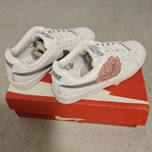 Nike Court Royale vit/silver med ros - Nike Court Royale sneakers i vitt skinn med silverfärgad swoosh och snörning. Ena sidan har en snygg rosa ros som detalj. Klassisk låg modell med rund tå och platt sula. Perfekta för dig som vill ha en stilren sneaker med unik touch. Storlek 42-43, helt nya. 