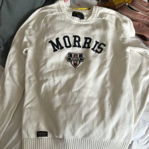 Vit stickad tröja från Morris - Säljer en vit stickad tröja från Morris med broderad logga och emblem framtill. Tröjan har rund hals, långa ärmar och ribbade muddar vid ärmslut och nederkant. Perfekt för dig som gillar klassisk och clean stil.