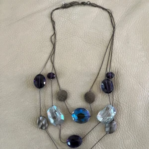 Långt halsband med blå och lila pärlor - Unikt långt halsband med flera lager tunna kedjor och stora, facetterade pärlor i blått, lila och transparent. Mellan pärlorna sitter runda, silvriga plattor med rutigt mönster. Perfekt för dig som vill sticka ut med coola detaljer och färg.