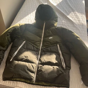 Grön pufferjacka från Nike Storm-FIT - Säljer en grön pufferjacka från Nike Storm-FIT med huva och vita dragkedjor, size M. Jackan har två stora fickor framtill och är perfekt för kalla dagar. Oversized vibe och riktigt skön att bära. Klassisk Nike-logga på bröstet.