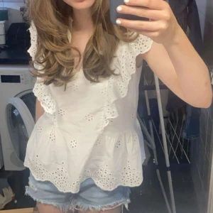 Vit blus med volang och broderi - Superfin vit blus med volangdetaljer och broderade hålmönster. Blusen är ärmlös och har en luftig passform med söt peplumkant. Perfekt för varma dagar och ger en romantisk vibe till din outfit.