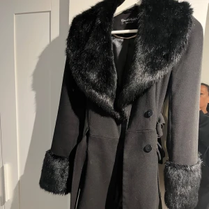 Svart kappa med fuskpäls från Miss Selfridge - Snygg svart kappa från Miss Selfridge med bred krage och muddar i fuskpäls. Dubbelknäppt framtill och med bälte i midjan💗 Perfekt under höst och vinter.