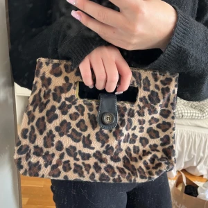 Leopardmönstrad kuvertväska med handtag - Säljer en kuvertväska med leopardmönster och inbyggt handtag. Väskan har en snygg svart läderdetalj med knäppning framtill. Materialet är mjukt och pälsliknande, perfekt för att sticka ut med en cool look.