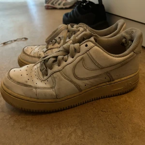 Nike Air Force 1 vita sneakers - Klassiska Nike Air Force 1 sneakers i vitt skinn med grå snörning och perforerad tå. Sulan är beige och har den ikoniska AIR-detaljen. Lågt skaft och rund tå, perfekt för dig som gillar streetstyle och tidlös design.