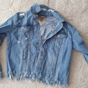 Blå jeansjacka med slitningar - Säljer en blå jeansjacka med snygga slitningar och fransad nederkant. Jackan har klassiska bröstfickor med knappar och långa ärmar. Perfekt för dig som gillar en avslappnad och trendig stil.