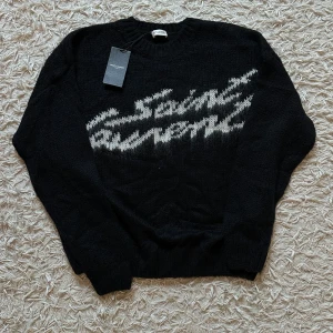 YSL- mohair sweater med lappar kvar  - Svart stickad tröja från Saint Laurent med vit, suddig logotyp framtill. Tröjan har rund halsringning och långa ärmar. Mjuk och fluffig känsla, perfekt för dig som gillar exklusiva statement-plagg. Denna versionen kallas för ”mohair sweater” och är helt perfekt plagg inför vintern så den håller en extremt varm medans den är jätteclean väldigt svår att få tag på och hög efterfrågan på tröjan. Tveka inte att skriva vid minsta funderingen! 