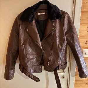 Brun skinnjacka från Gina Tricot - Snygg brun skinnjacka från Gina Tricot med svart fuskpäls på kragen. Jackan har klassisk bikerstil med dragkedjor, knappar och bälte i midjan. 