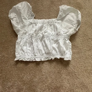 Vit blus med puffärmar från Zara - Supersöt vit blus från Zara med puffiga korta ärmar och volangdetaljer. Blusen har smockad resår vid hals, midja och nederkant som ger en snygg croppad look. Perfekt för dig som gillar romantisk stil och vill ha något luftigt till sommaren.