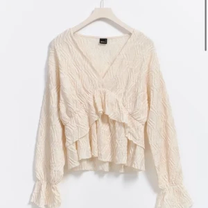 Vit blus med volang och struktur - Supersöt vit blus med v-ringning och lång ärm. Blusen har en unik strukturerad yta och en snygg volangdetalj framtill som ger extra stil. Perfekt för dig som gillar romantiska och trendiga plagg med det lilla extra.